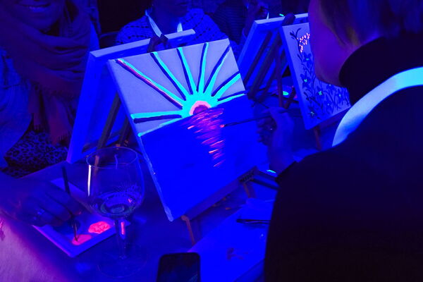 Malende Personen mit kleinen Keilrahmen und Neonfarben unter Schwarzlicht.