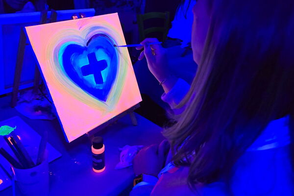 Malende Person mit kleinem Keilrahmen und Neonfarben an Tisch unter Schwarzlicht.