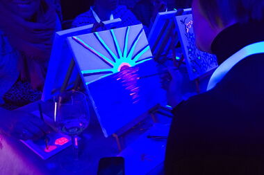 Malende Personen mit kleinen Keilrahmen und Neonfarben unter Schwarzlicht.